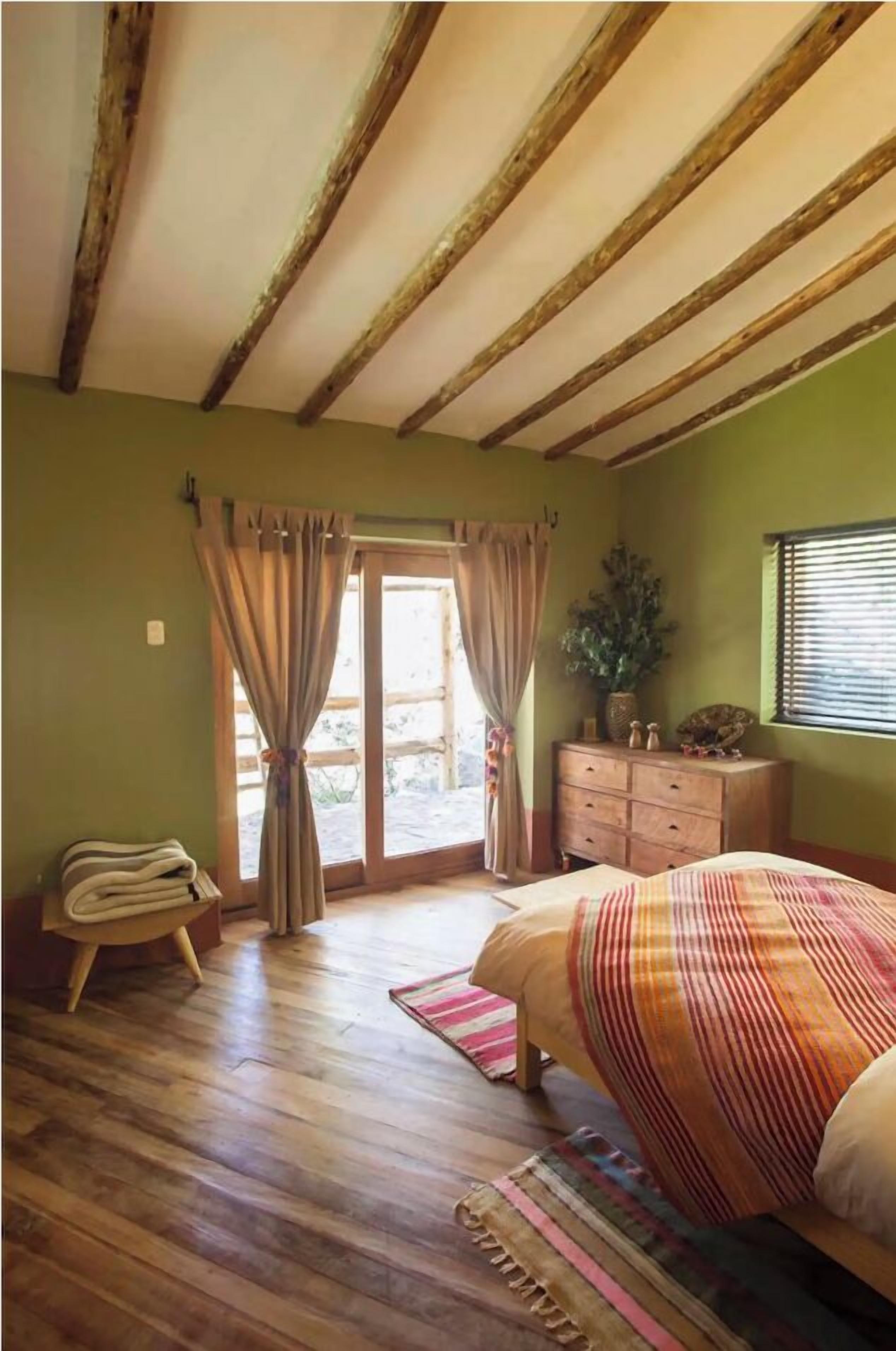 Une chambre avec des poutres en bois, un lit recouvert d’une couverture à rayures, une commode, une plante et une fenêtre avec des stores.