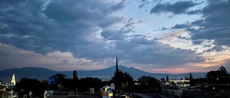 A cityscape at dusk with a prominent mountain in the background, a church spire, and a small building with a yellow-lit window.