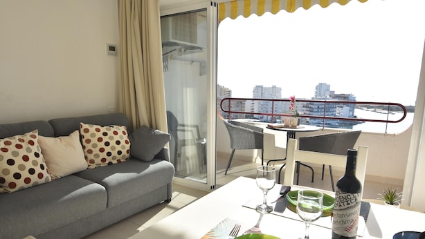 Un salon moderne avec un canapé gris, une table à manger agrémentée d’une bouteille de vin, et une vue sur la ville depuis un balcon.