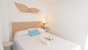 Un lit bien fait, recouvert d’une couvre-lit blanche, avec un oreiller à motifs bleus et une serviette ornée de fleurs. Une table de chevet avec une lampe se trouve à côté du lit, et une étagère est fixée au mur au-dessus du lit.