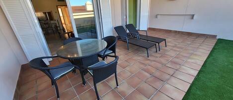 An outdoor patio with a round glass table and four chairs, surrounded by a tiled floor and a small patch of artificial grass.
