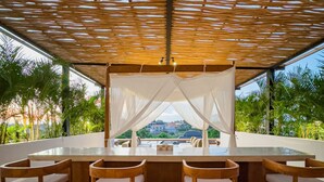 An outdoor bar area with a wooden table and chairs, a canopy overhead, and a view of greenery and buildings in the background. - Narawangsa Villa | 4br | Rooftop West Coast View (Kerobokan)