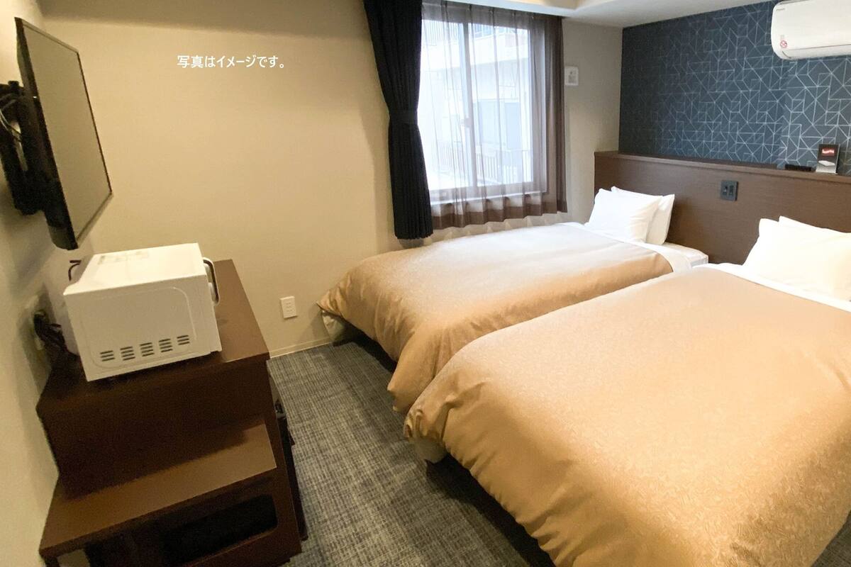 Habitación de hotel con dos camas, un escritorio con microondas, una ventana con cortinas y aire acondicionado de pared.