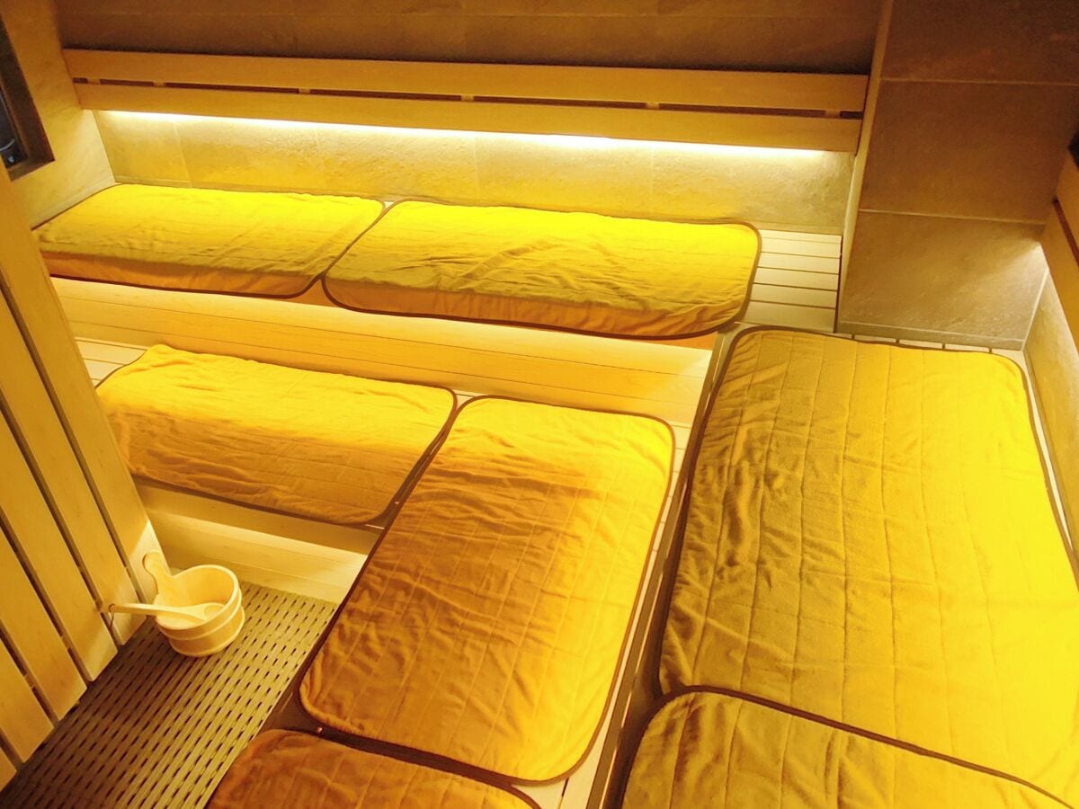 Quarto pequeno com duas camas, cada uma com colchão e almofadas amarelas combinando. Há um banquinho pequeno e uma bacia branca no chão.