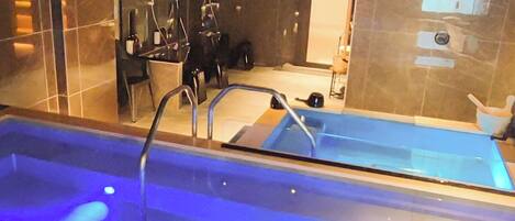 A modern indoor swimming pool with a glass barrier, a metal ladder, and a blue-lit water feature.