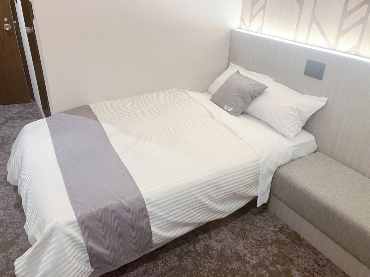 A neatly made bed with a grey and white bedspread, a grey pillow, and a headboard with a light switch.