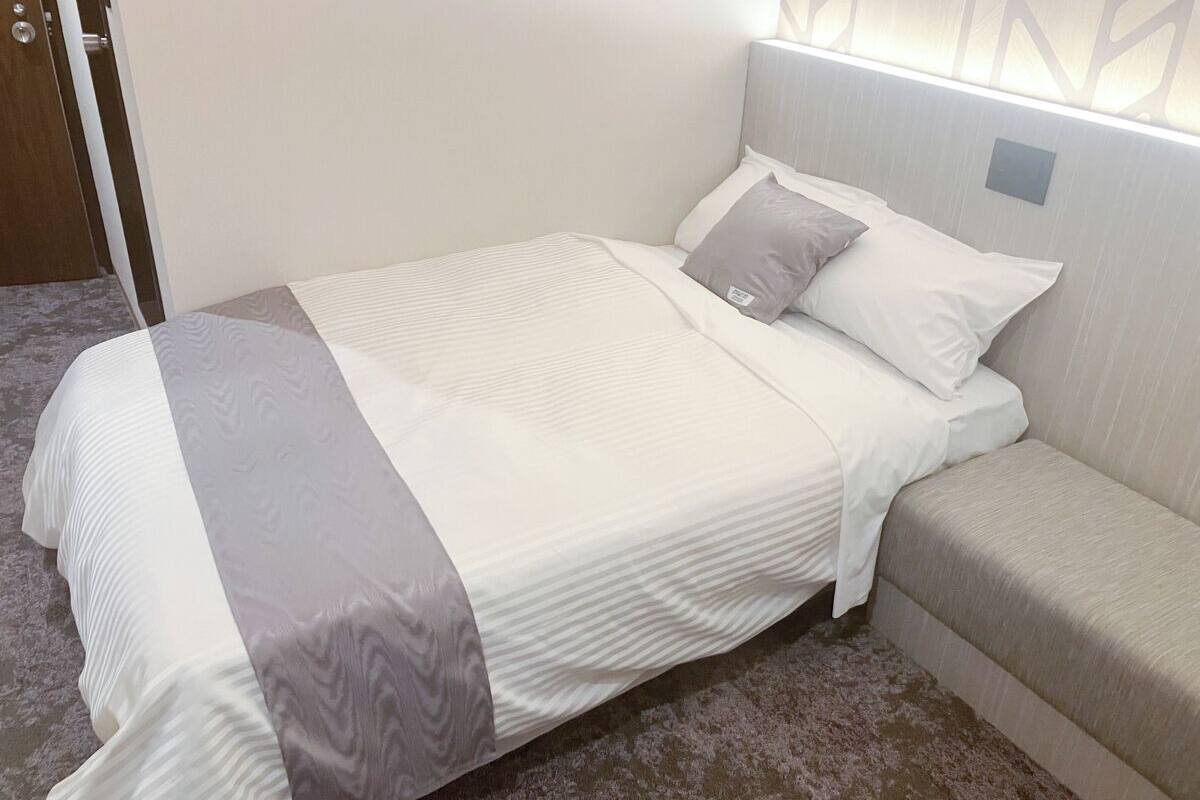 Una cama bien hecha con una colcha gris y blanca, una almohada gris y un cabecero con un interruptor de luz.