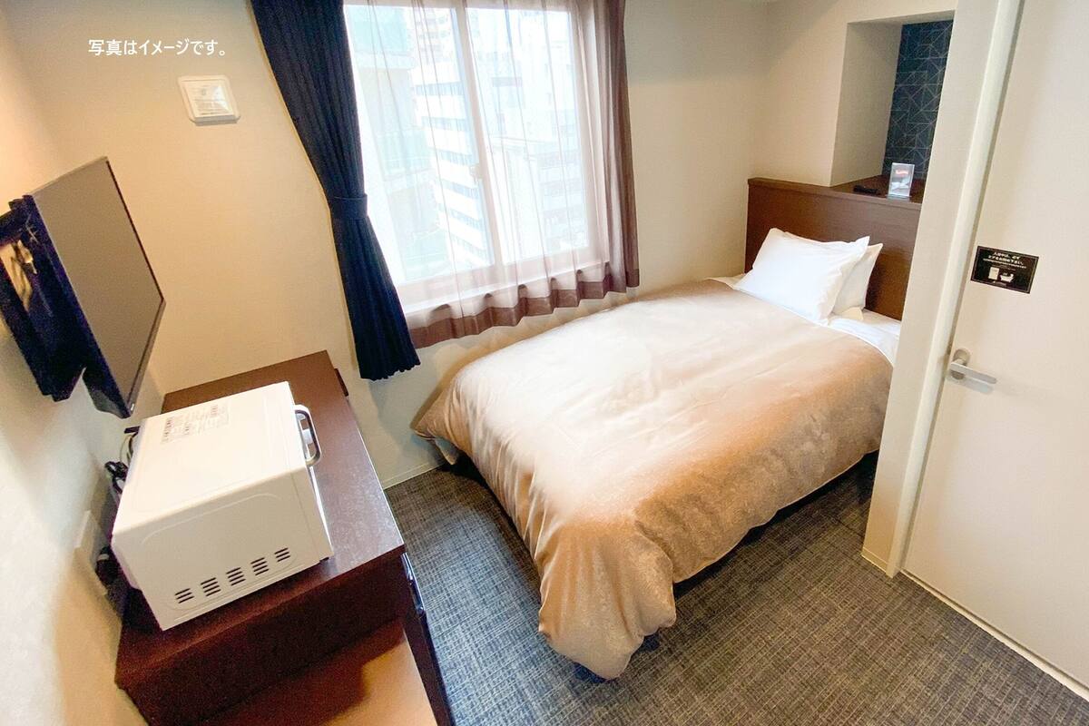 Una habitación de hotel con cama, un escritorio con un electrodoméstico blanco, una ventana con cortinas y una puerta con un letrero.