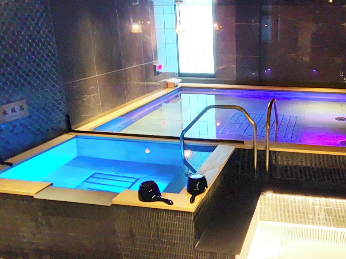 Une piscine intérieure moderne avec un fond en verre, deux bouilloires noires posées sur le bord et une échelle métallique menant à l’intérieur de la piscine.