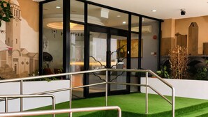 Un hall d’hôtel doté d’une façade en verre, d’un escalier avec des rampes en métal et d’un espace recouvert de gazon synthétique vert.