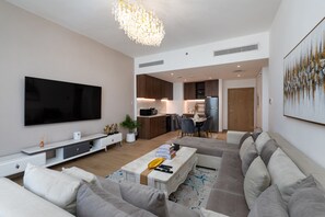 A modern living room with a large flat-screen TV, a sofa, a coffee table, and a dining area with a kitchen in the background. - Contemporary La Mer 1BR w/ Beach Access (Dubai)