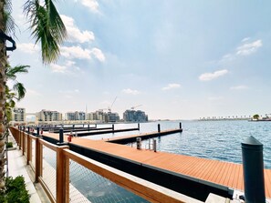 A waterfront promenade with a wooden deck, a chain-link fence, and a view of modern buildings and a bridge in the distance.