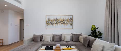 Un salon moderne avec un canapé d’angle, une table basse sur laquelle on trouve des livres et une banane, et un grand tableau abstrait accroché au mur.