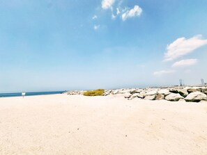 A beach with a rocky shoreline, a clear sky, and distant construction cranes.
