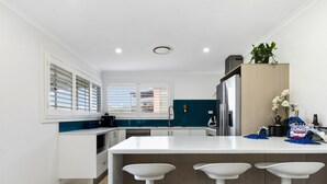 Una cocina moderna con una isla central, taburetes blancos, un refrigerador de acero inoxidable y una ventana con persianas.