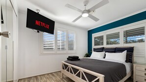Un dormitorio con cama, televisor montado en la pared, ventilador de techo y ventana con persianas.