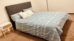 Un lit bien fait, avec une couette à motifs gris et blancs, deux oreillers et une tête de lit sombre. Une petite table de chevet blanche, sur laquelle se trouve un panier en osier, est visible sur la gauche.