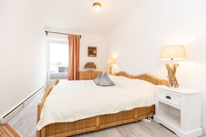 A bedroom with a bed, bedside tables, a lamp, a picture frame, and a window with curtains. - Jamé - Bright 2-room apartment with balcony, parking space and WLAN in Westerland. (Westerland)