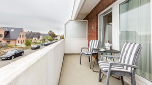 A balcony with two chairs and a table, a brick wall, and a view of parked cars and houses. - Jamé - Bright 2-room apartment with balcony, parking space and WLAN in Westerland. (Westerland)