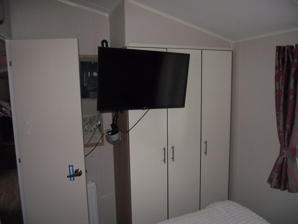 A bedroom with a bed, a wardrobe, a television mounted on the wall, and a door with a blue arrow sign. - Seafront Caravan - Golden Sands - 6 Berth by Your Away Stays (Rhyl)