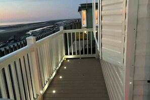 A balcony with a wooden floor and white railings, overlooking a beach and ocean. - Seafront Caravan - Golden Sands - 6 Berth by Your Away Stays (Rhyl)