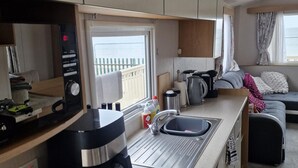 Een kleine keuken met een koffiezetapparaat, een waterkoker en een spoelbak. Er is een raam met uitzicht op een balkon en een bank met een plaid.