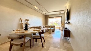 A modern living room with a dining area, a sofa set, a flat-screen TV mounted on the wall, and a view through a large window. - Marrakech Prestigia Piscine Golf Saphir (Marrakech)