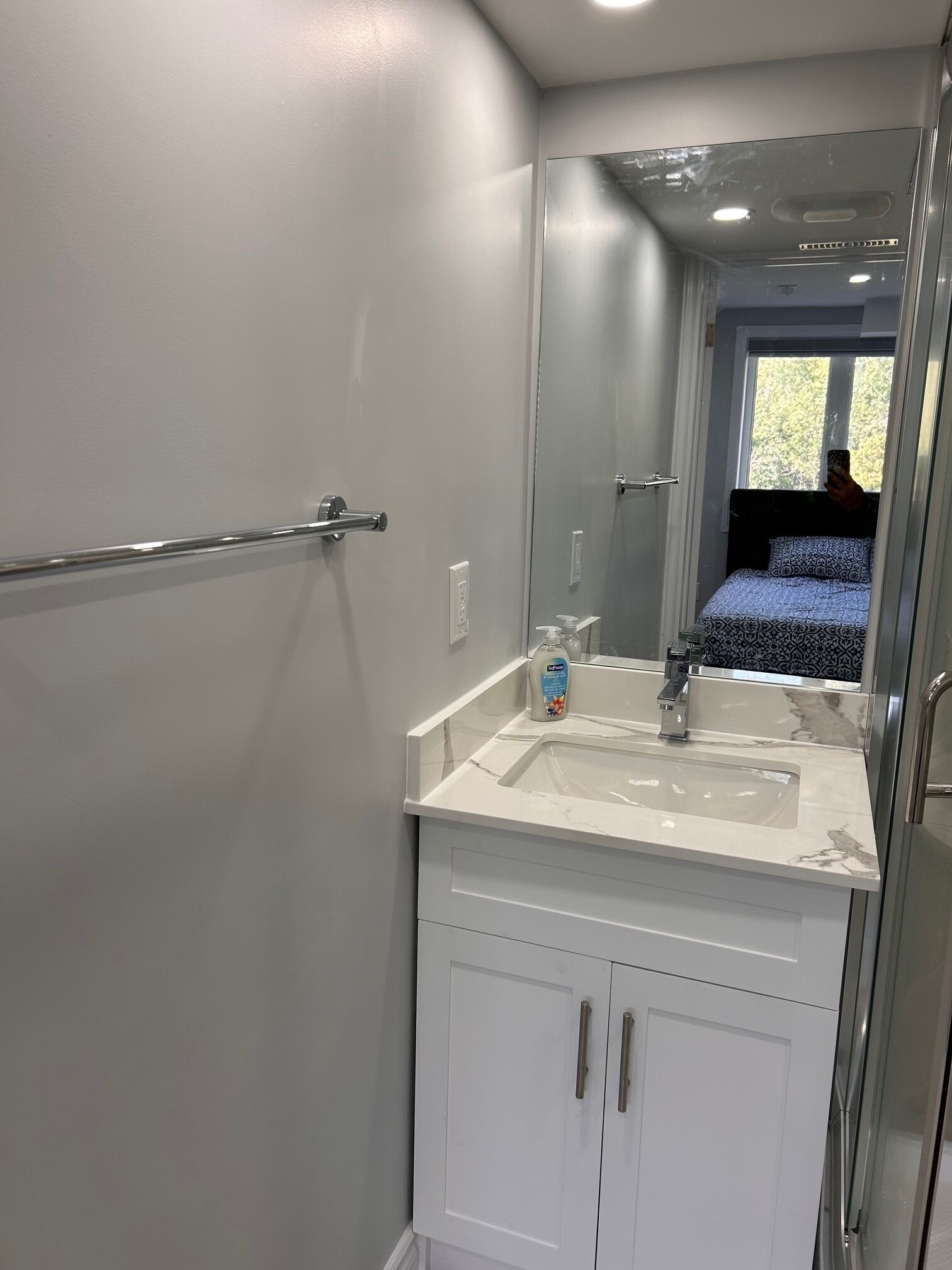 A bathroom with a white vanity, a mirror, a hand soap dispenser, and a window with a view of greenery.