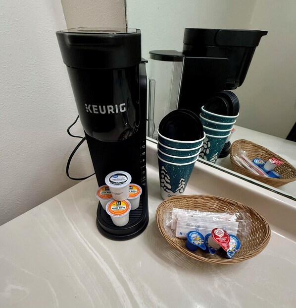 A Keurig coffee maker with a drip tray, a basket of coffee pods, and a stack of disposable cups.