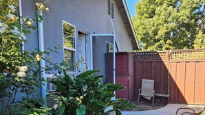 A residential backyard with a concrete pathway leading to a house with a blue exterior. There is a wooden fence, a bench, and various plants and flowers. - Charming 1-bedroom house in superb San Jose with AC, WiFi (San Jose)