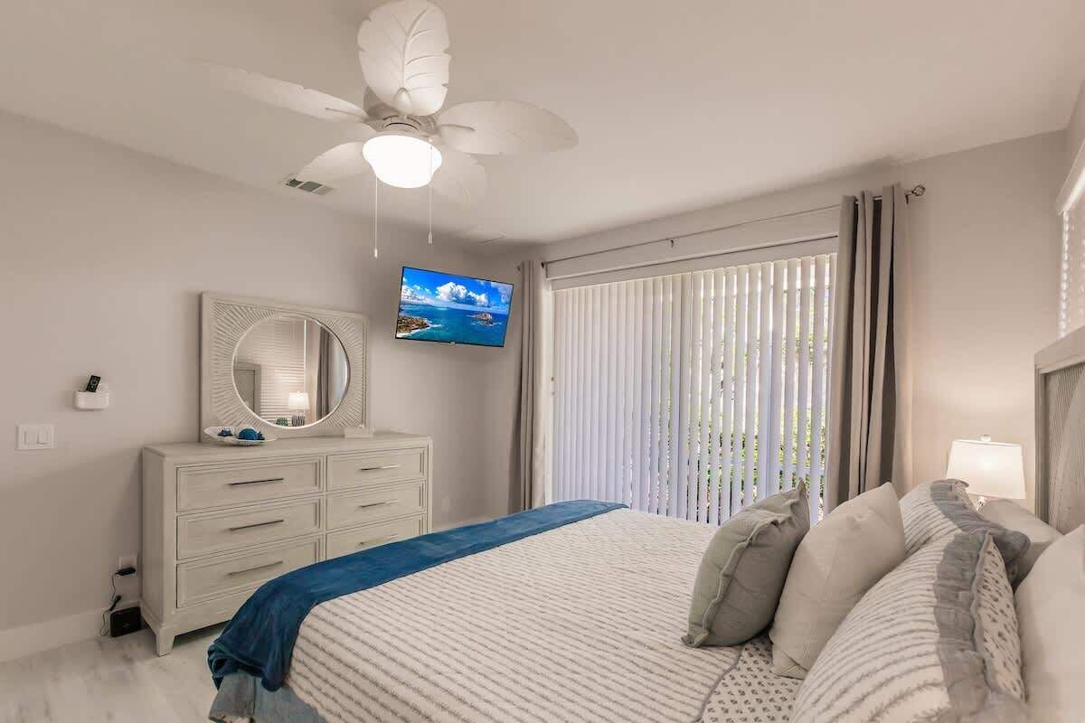Een slaapkamer met een bed, een plafondventilator, een televisie, een dressoir en een raam met jaloezieën.