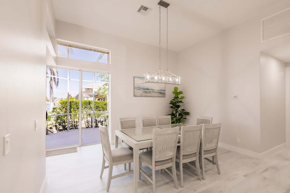 A dining area with a white table and chairs, a hanging light fixture, and a view of greenery outside.