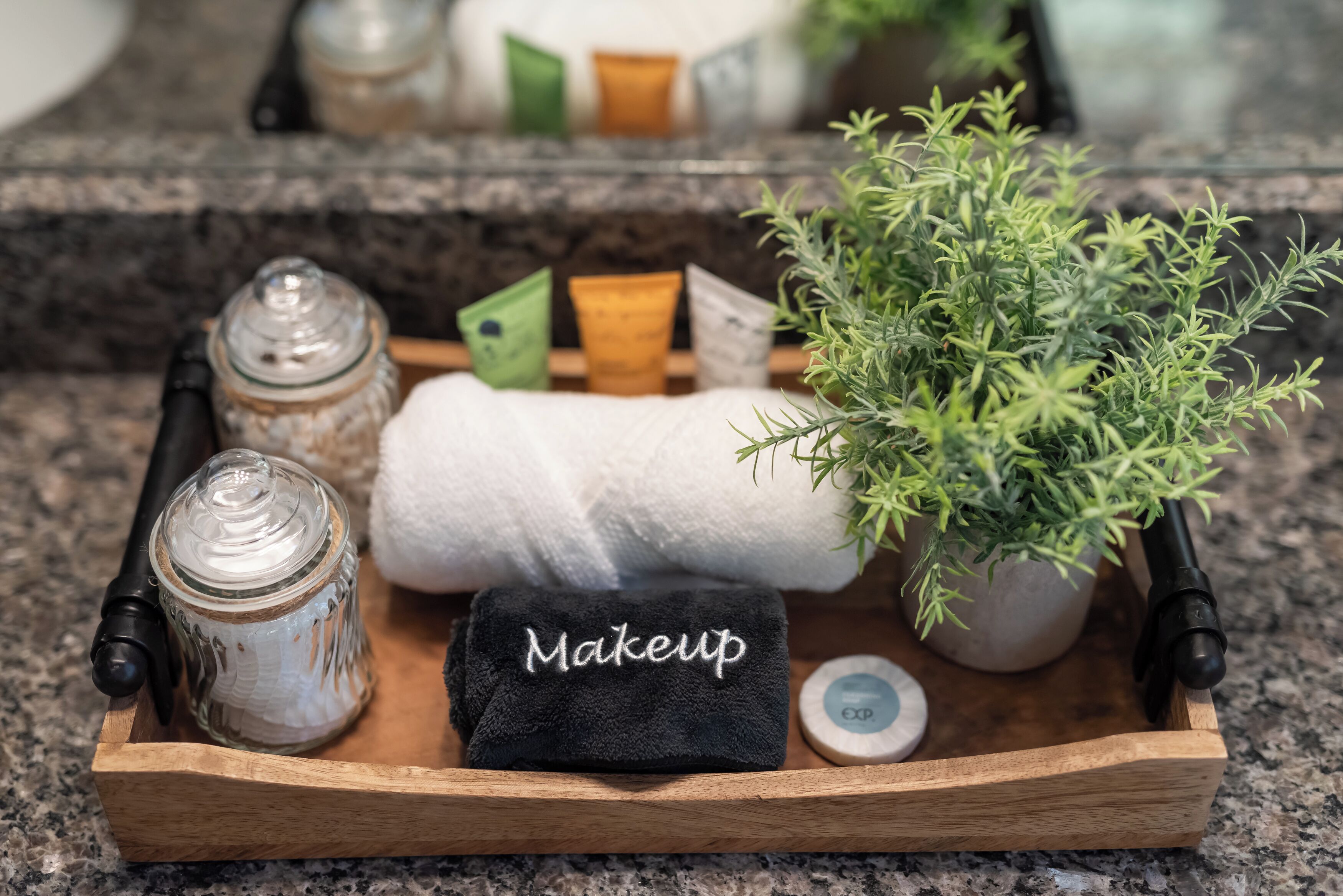 Un plateau en bois contenant une trousse de maquillage, une plante en pot, un bocal et un récipient, le tout posé sur un comptoir en granit.
