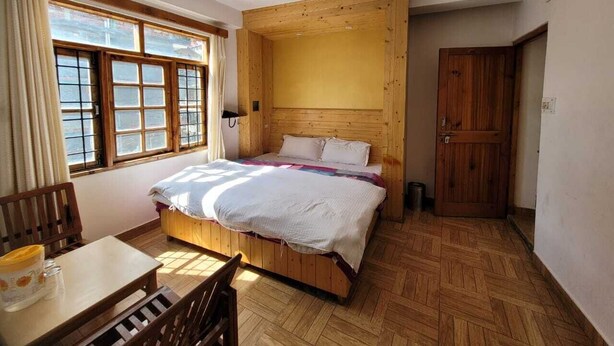 A bedroom with a wooden headboard, a bed with white bedding, a wooden table with chairs, and a window with curtains. - Hills Homestay and Devallya (Manali)