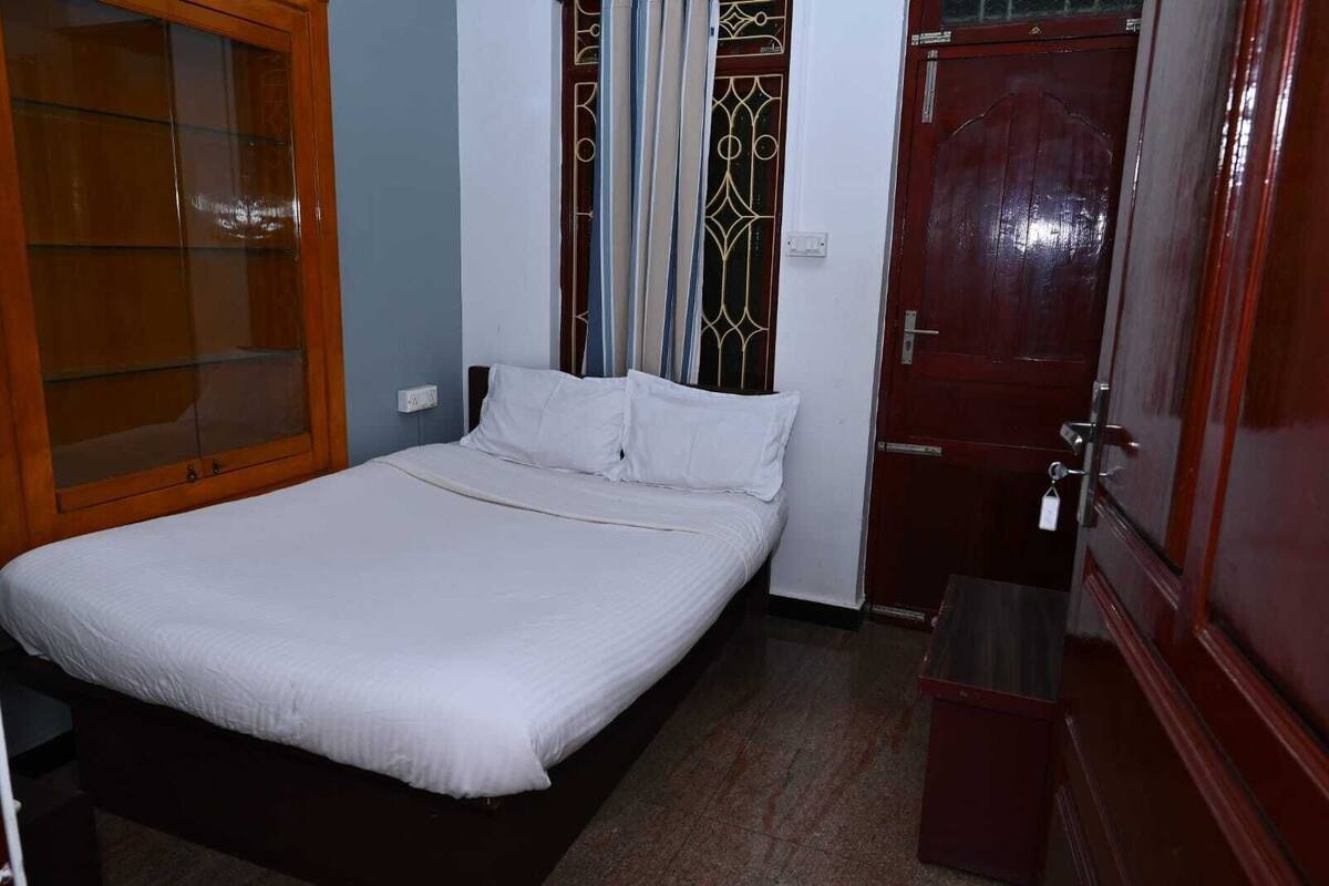 A single bed with white bedding in a room with a wooden wardrobe, a door with a metal handle, and a small trash bin.