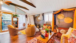 A cozy living room with a wooden floor, wicker furniture, a colorful rug, and a large window with curtains.