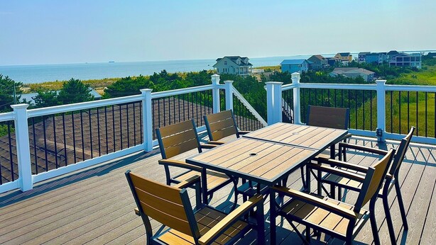 A wooden deck with a table and chairs, overlooking a beach and houses.