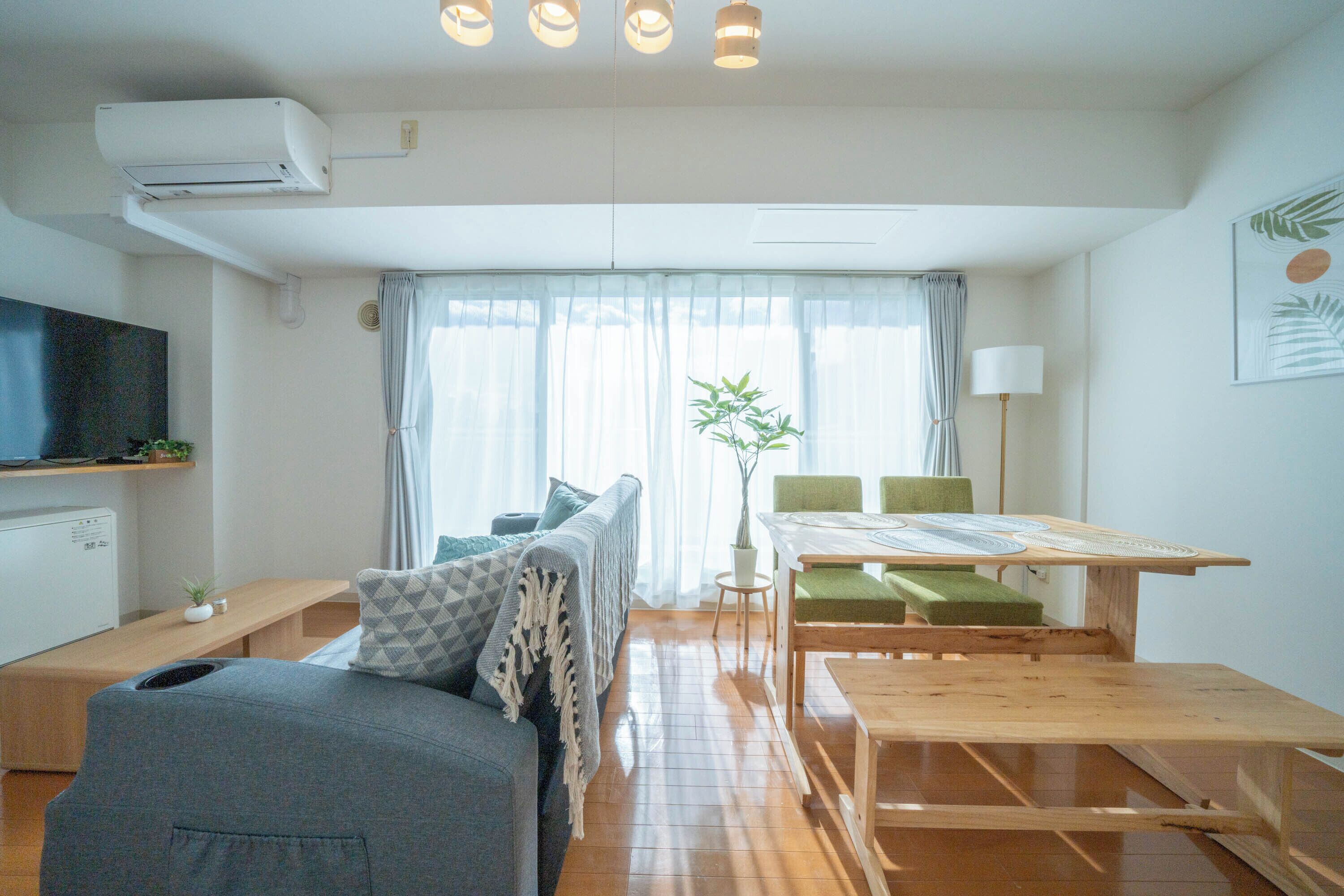 A modern living room with a sofa, a dining table with chairs, a TV mounted on the wall, and a window with curtains.