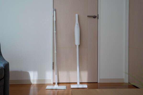 A room with a wooden floor, a grey sofa, and two white upright vacuum cleaners. - Sapporo irios cozy inn (Sapporo)