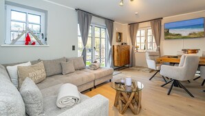 A modern living room with a grey sofa, a glass coffee table, and a dining area with a wooden table and chairs. - Haus Rosi - Holiday home in Westerland with sauna and garden. Pets are (Sylt)
