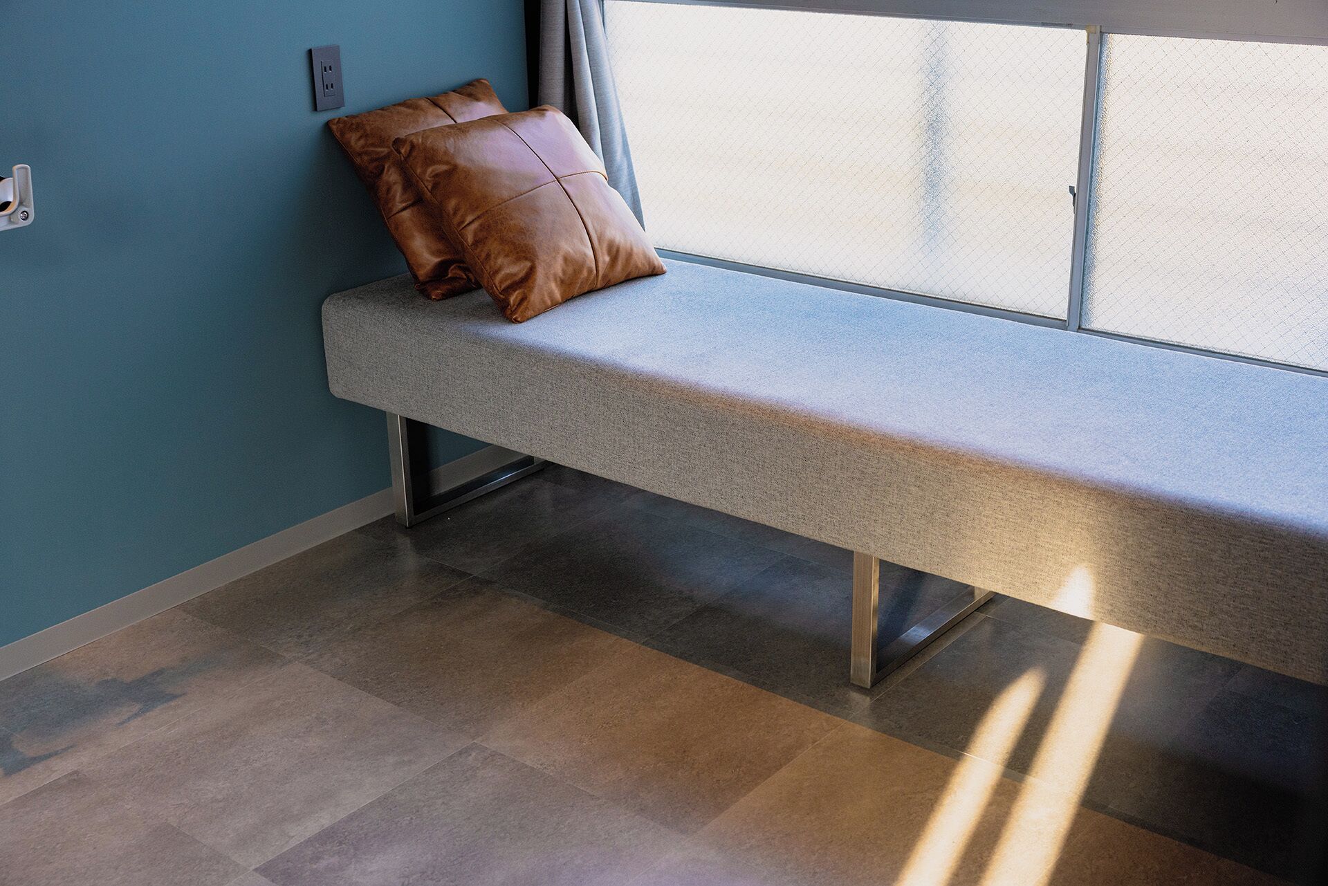 A bench with a brown cushion against a wall with a window.