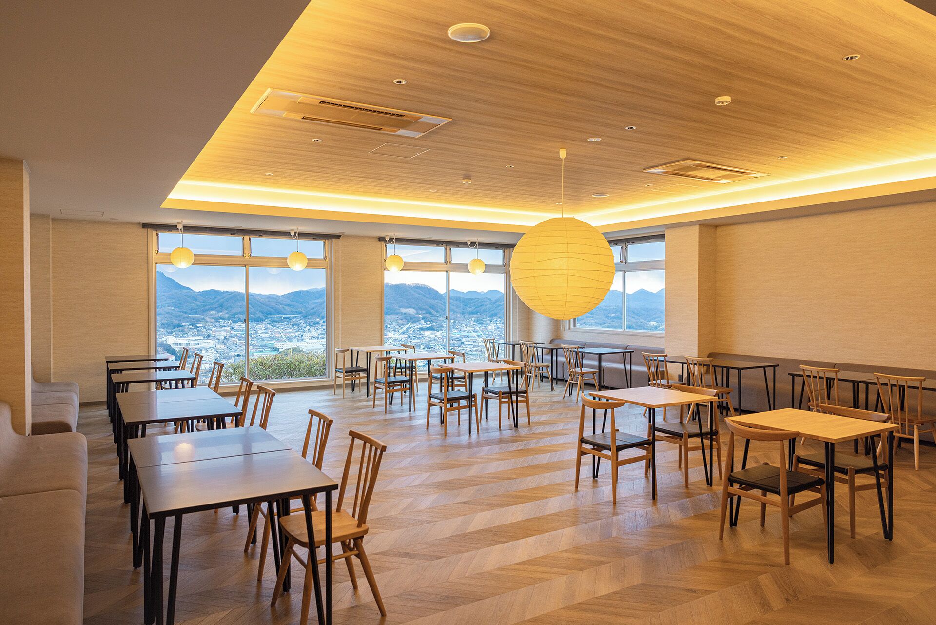 A spacious dining area with multiple tables and chairs, large windows offering a view of a city and mountains, and a wooden ceiling with recessed lighting.