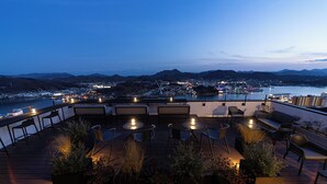 A rooftop terrace with seating and potted plants, overlooking a cityscape and a body of water at dusk. - onomichi club (Onomichi)