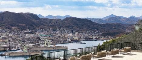 A coastal cityscape with a harbor, mountains in the background, and a balcony with wicker furniture.