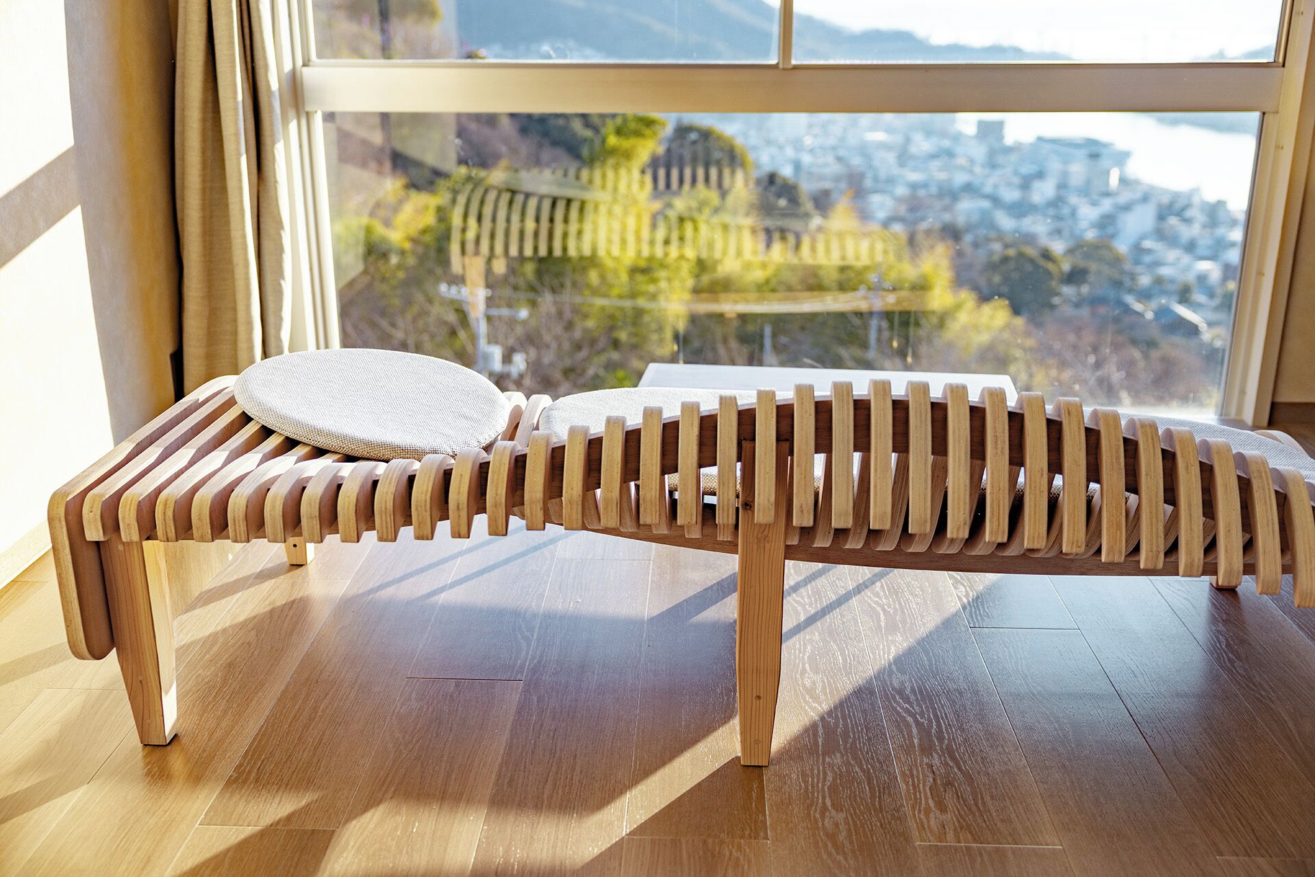 A wooden lounge chair with a woven seat cushion, positioned on a wooden floor near a large window with a view of a cityscape and mountains.