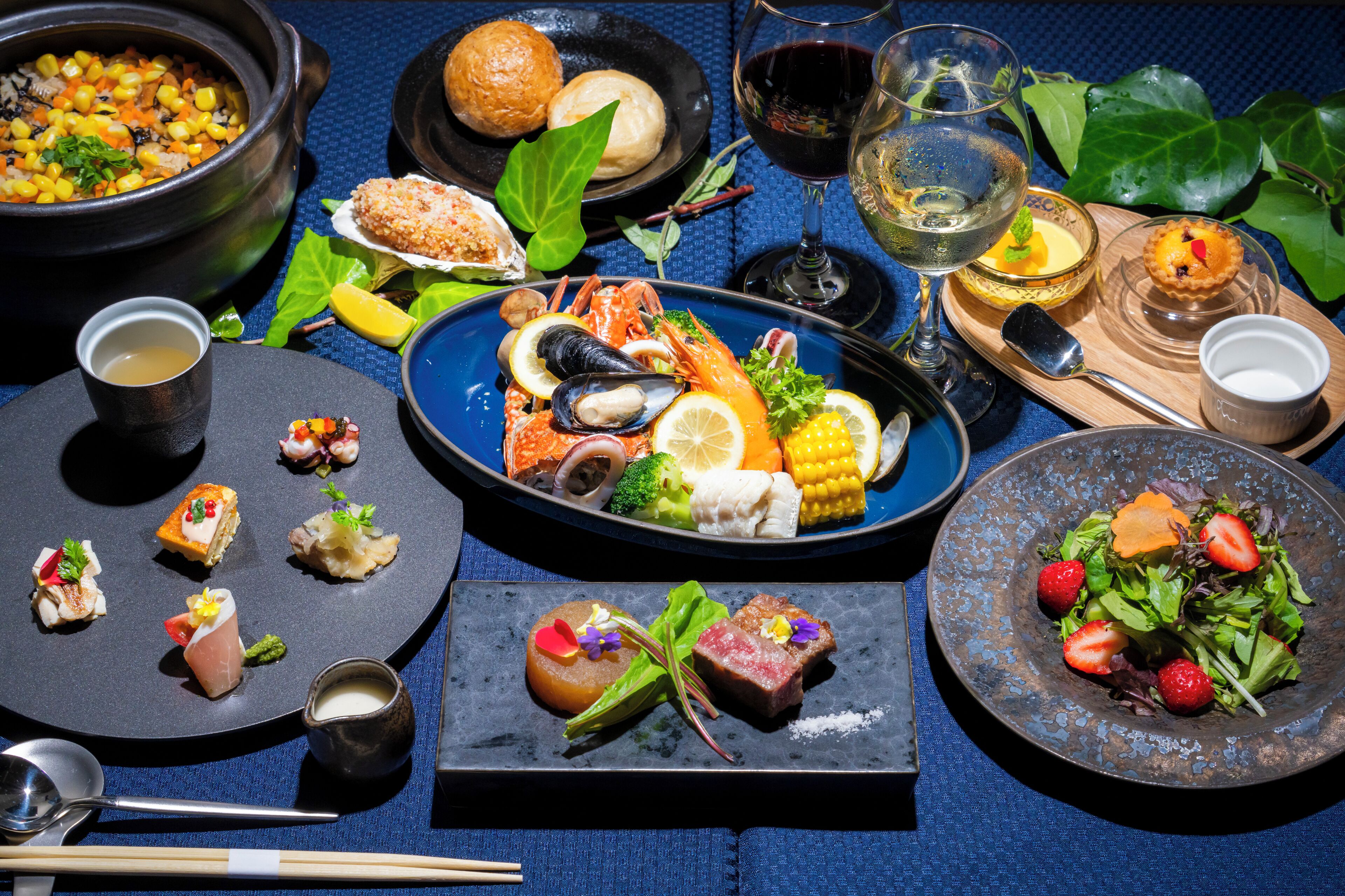 Una variedad de platos que incluyen mariscos, verduras y postres, dispuestos sobre una mesa con mantel azul.