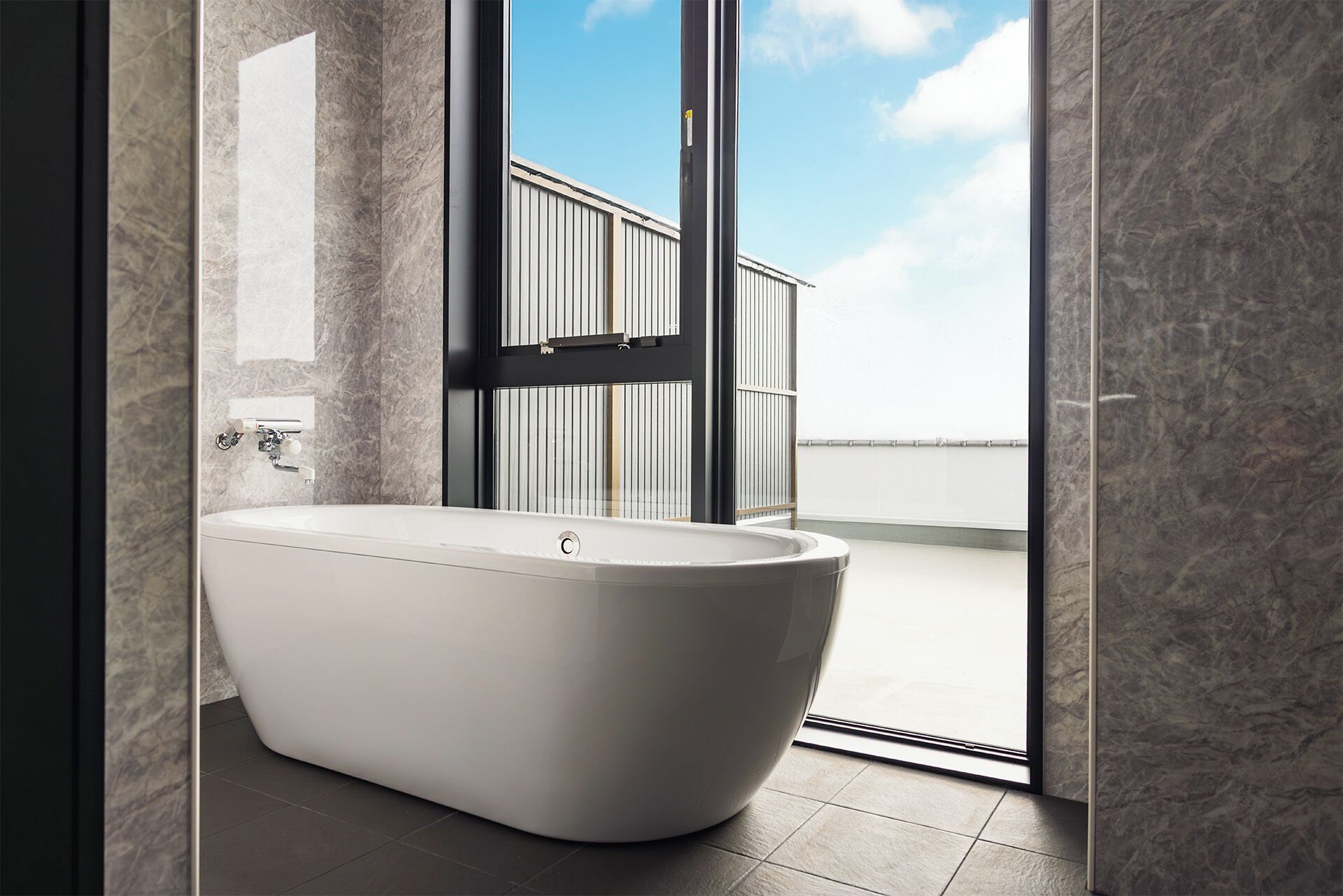 Een moderne badkamer met een vrijstaand bad, een glazen deur naar een balkon, en uitzicht op een gebouw aan het water.