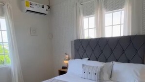 Un dormitorio con una cama grande, ropa de cama blanca, un cabecero gris y una ventana con cortinas.