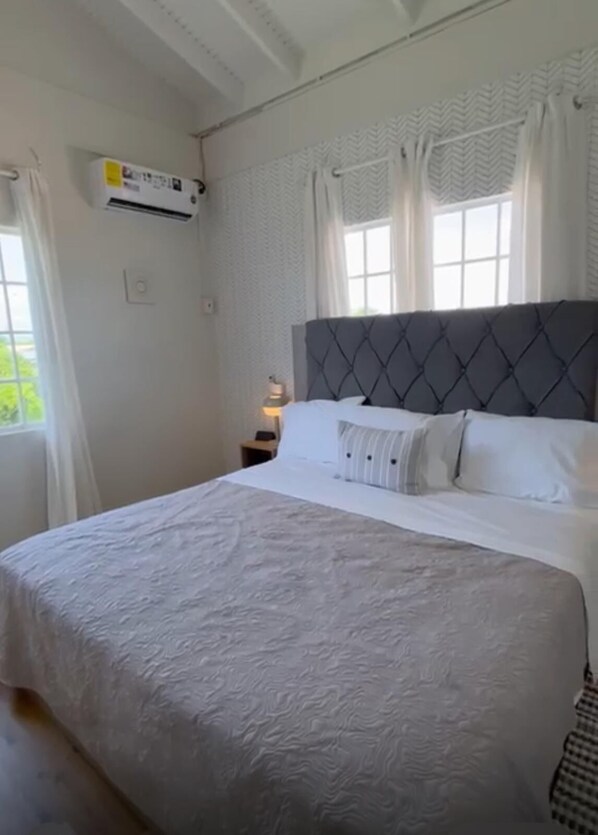 Ein Schlafzimmer mit einem großen Bett, weißer Bettwäsche, einem grauen Kopfteil und einem Fenster mit Vorhängen.