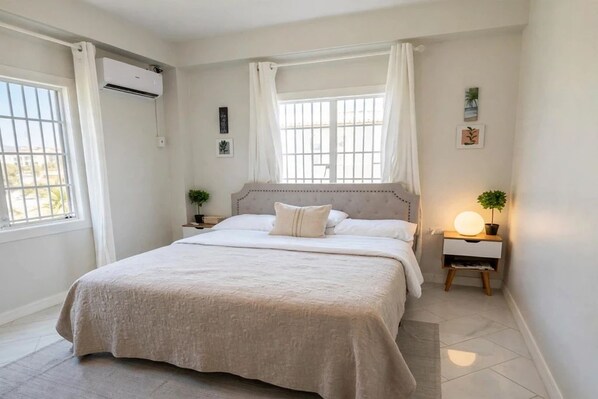 Un dormitorio con una cama grande, una mesita de noche con lámpara y un ventanal con cortinas.